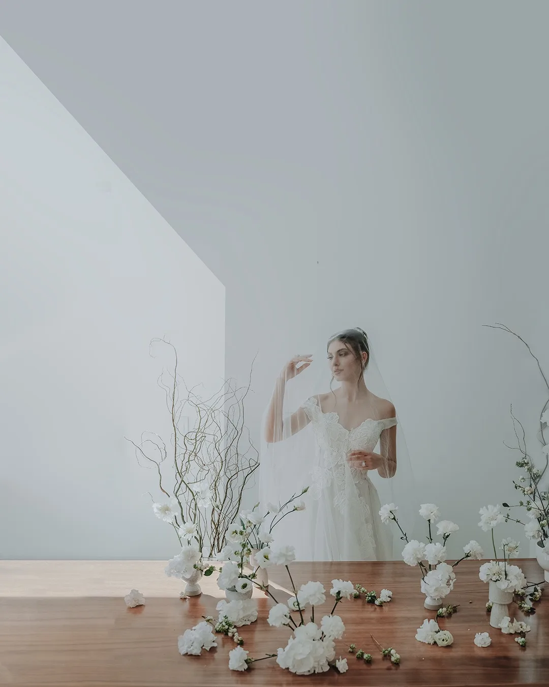 Minimalist Wedding Floral Arrangement with Bride – Floba Flower Boutique Elegant bride in an off-shoulder lace wedding dress with a modern minimalist white floral arrangement on a wooden table, styled by Floba Flower Boutique in Vancouver.