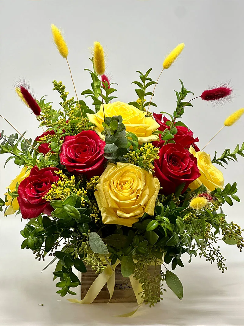 Floral arrangement of red and yellow roses with green foliage and decorative accents in a wooden box, named Sunset Romance.